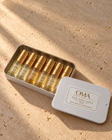 The OMA Edit SIX open white keepsake tin with six labeled perfume vials and gold caps on warm ivory stone; example scent selection