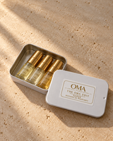 The OMA Edit THREE open white keepsake tin with three labeled perfume vials and gold caps on warm ivory stone; example scent selection